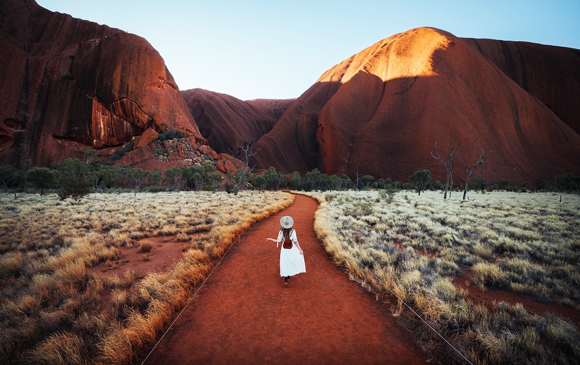 a woman in a white dress walks up to Uluru in the early morning.